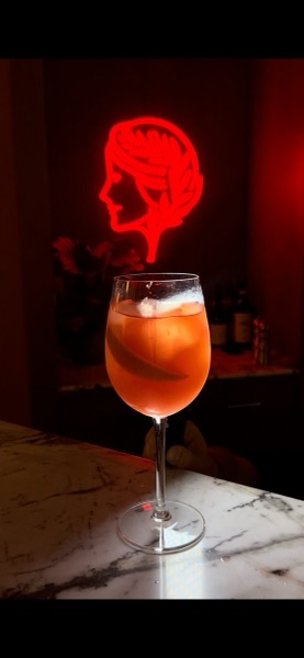 Ein Spritz auf dem Marmortresen vor einem roten Neonkopf im Profil.