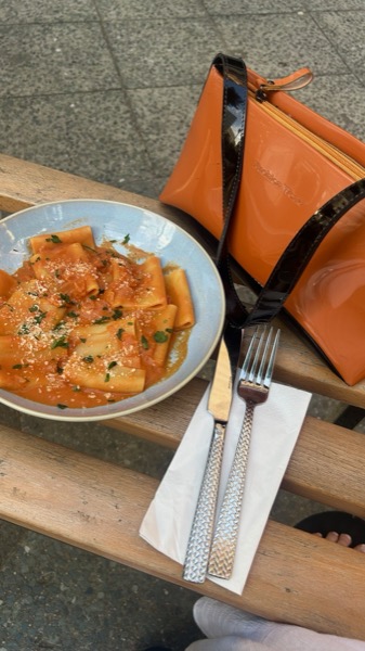 Ein Teller Paccheri mit Tomatensauce auf einer Holzbank, daneben Besteck und eine Tasche.