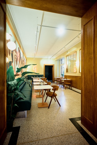 Der lange Gastraum bei Tageslicht: Terrazzoboden, grüne Bänke, eingedeckte Tische bis zum Fenster.