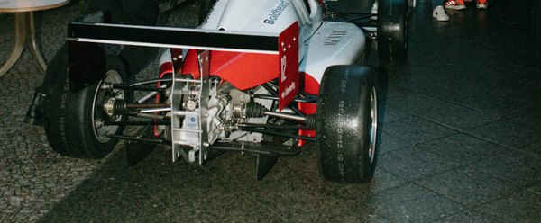 Ein weiß-roter Formel-Rennwagen mit der Startnummer 12 steht abends auf dem Kopfsteinpflaster vor der Bar, Heckflügel und Diffusor zur Kamera.